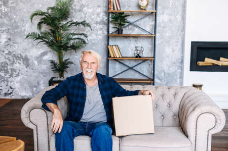 Satisfied mature man received a long-awaited shipping. A senior gray-haired man sits on sofa with cardboard box, looks at camera and smiles cheerfullyの写真素材
