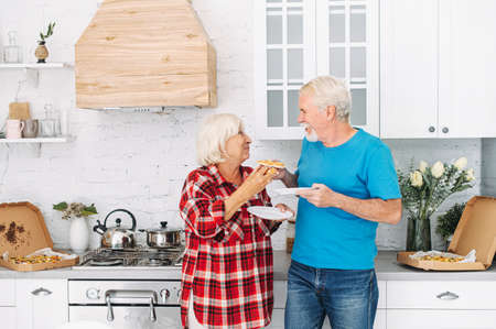 Cheerful senior couple in love eating pizza together in the kitchen at homeの写真素材