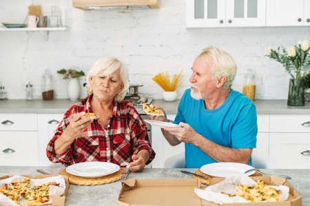 Senior couple in love eating pizza together in the kitchen at home. Beautiful elderly woman and grey-haired mature man are biting pieces of pizzaの写真素材