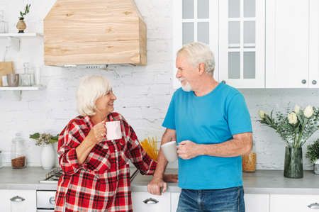 Loving senior spouses drink tea in the kitchen at home. A mature husband and beautiful grey-haired wife with cups in the hands looks to each other with love and careの写真素材