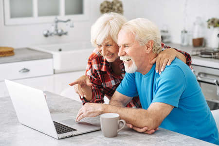 Cheerful senior couple looks with interes on laptop screen. Grey-haired smiled spouses make shopping togetherの写真素材