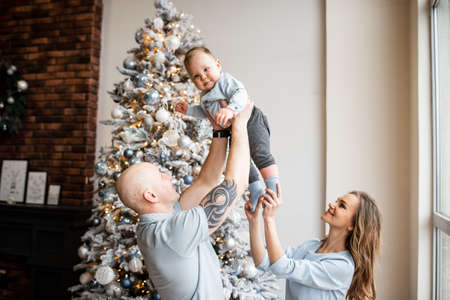 Happy family in cozy living room with decorated Christmas tree. Young father is throwing his little son up, a beautiful mother is laughingの写真素材