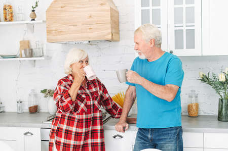 Loving senior spouses drink tea in the kitchen at home. A mature husband and beautiful grey-haired wife with cups in the hands looks to each other with love and careの写真素材