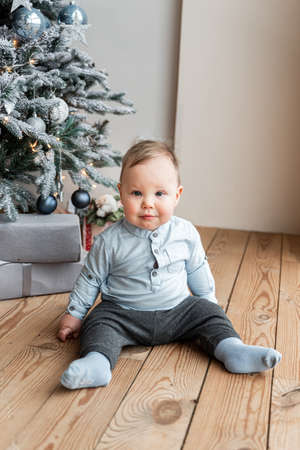 Funny cute baby boy in blue clothes sits indoors on the floor near festive Christmas tree and smilesの写真素材