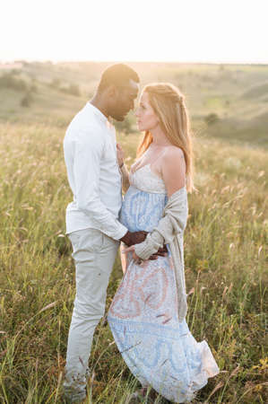 Stylish diverse couple. An african-american husband and his caucasian pregnant wife are holding hands and hugging outdoorsの写真素材