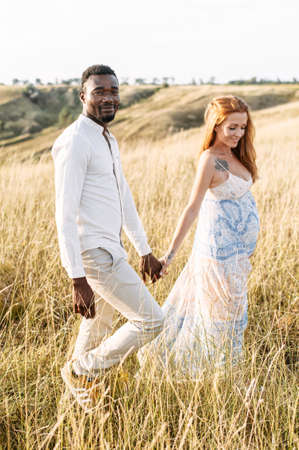 Romantic couple in love walking on the meadow. A beautiful pregnant red haired wife and her handsome black husband hugging outdoorsの写真素材