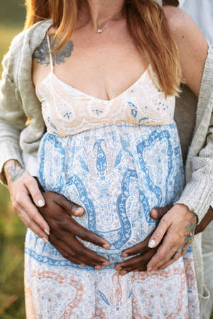 Hands of an african american father and white mother on the pregnant belly outdoor, faces are not...