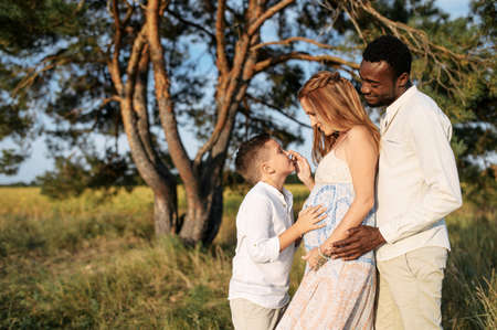Happy diverse family spends time outdoors together. Pregnant wife, care husband and school age boy on the meadow in warm summer dayの写真素材