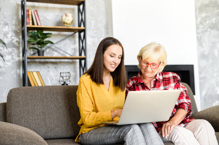 Senior mother and adult daugther sit on the sofa at home and look with interest on computer screen. Teaching the older generation to use a laptopの写真素材