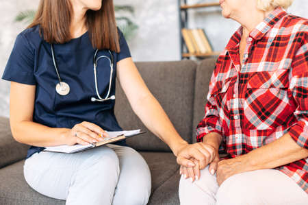 Young nurse talking with elderly patient at home care medical visit. Doctor holds hand of senior woman with empathy and careの写真素材