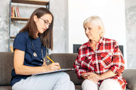 The doctor came to the patient's call. A senior woman complains to the doctor or nurse and tells the symptoms sitting on the sofa at homeの写真素材