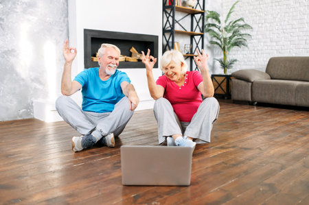 Elderly couple practicing yoga with online video classes. A grey-haired man and woman sits on the floor in lotus pose and look at laptop screenの写真素材