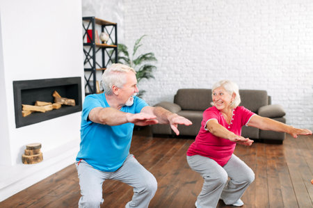 Elderly couple doing squats together at home. Cheerful grey-haired spouses doing exercises, look to each other and smilesの写真素材