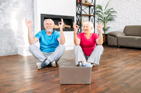 Elderly couple practicing yoga with online video classes. A grey-haired man and woman sits on the floor in lotus pose in front of laptopの写真素材