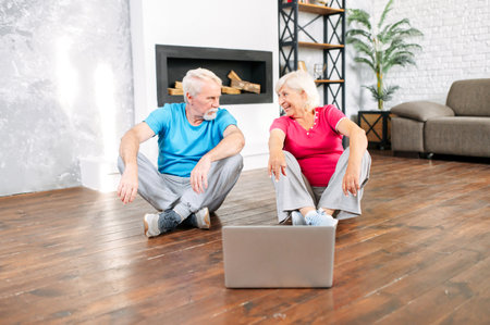 Senior couple going to practicing yoga at home with an online video classes. Elderly spouses in sportswear look to each other sitting on the floor at home in front of the laptopの写真素材