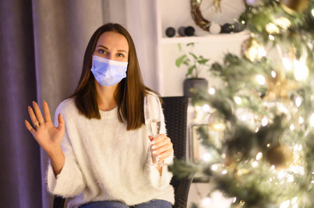 A woman wearing protective medical mask having video call at the Christmas eve. Webcam shot of a girl with a glass of champagne near festive Christmas tree during pandemicの写真素材