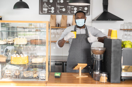 Friendly and cheerful african-american male waiter, barista in apron and protective mask holds paper cup with hot coffee and shows thumb up standing behind counter in modern loft coffee shop, bakeryの写真素材