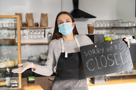 Cafe, bar closed due outbreak lockdown, female wearing medical mask and apron looks at camera, holds a closed sign and shrugs shoulders. Small business crisis conceptの写真素材