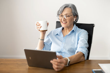 Busy overloaded with work senior female businesswoman talking to family on a video-call, using a tablet, sitting at the desk at home, in the office, wearing glasses, earphones, and Fitbitの写真素材