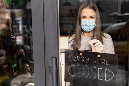 View through glass door female wearing medical mask and an apron holds closed sign board. Small...