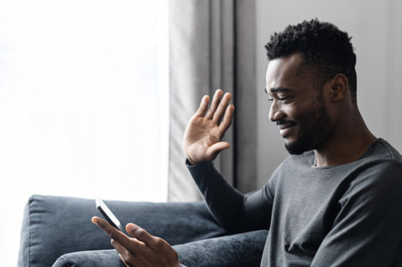 Side view an African guy is talking online via video call on the smartphone sitting at home on the couch, video conference with family, employees, friends, virtual meetingの写真素材
