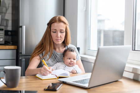 Focused young woman holds infant in arms and making notes. A busy mother babysit adorable little daughter while studying from home. Female student watching online lecture, education conceptの写真素材