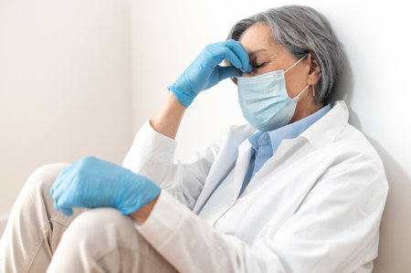 Stressed and tired sad female doctor in uniform, face mask and gloves leaning on the wall in despair. Upset nurse resting, taking a break. Rest at work during a pandemic of coronavirus conceptの写真素材