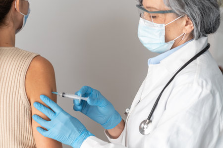 Portrait of two people, senior female medical professional wearing gloves and face mask injecting a vaccine to a young lady, a girl is getting a shot of a flu vaccine, hospital healthcare caseの写真素材