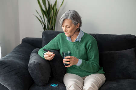 Senior mature gray-haired lady sitting in the living room on the couch or sofa, holding one of the blue capsules from a blister pack of prescribed medicine and a glass of waterの写真素材