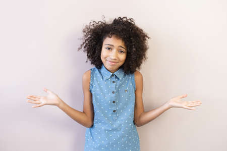 Portrait of cute biracial preteen girl shrugs shoulders, a schoolgirl wearing polka dot shirt looks at camera spreads her hands as a sign of misunderstanding, incomprehension, isolated on pastel pinkの写真素材