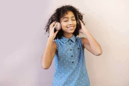 Cute African preteen girl and enjoying listening music in the headphones, the schoolgirl dreaming with eyes closed on a pastel pink backgroundの写真素材