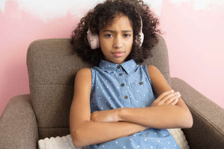A curly African-American school-age girl wearing headphones sits on the armchair frowning face in displeasure, keeps arms folded. Difficult teen-ageの写真素材
