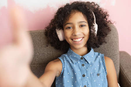 A cheerful curly African-American preteen girl wearing a headphones is taking a selfie. A close-up...