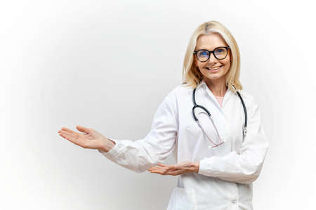 A charming female doctor pediatric, physician, therapist wearing a white medical gown with a stethoscope on the shoulders points hands on empty space, mock up, copy space. Healthcare and medicineの写真素材