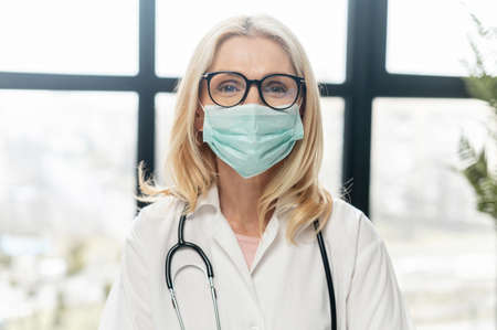 A close-up of a female doctor wearing protective face mask and a medical robe with a stethoscope on the shoulders, portrait of a middle-aged healthcare professional working during pandemicの写真素材