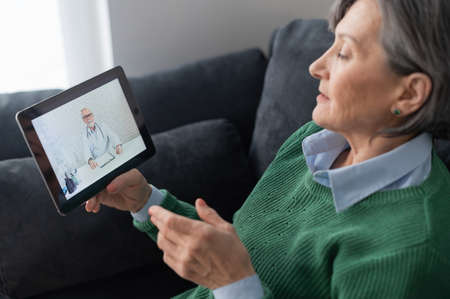Mature senior lady holding a tablet or pad and talking to a doctor online from home, having a virtual medical check-up with the medical specialist on a webcam. Cyber medicine and stay at home conceptsの写真素材