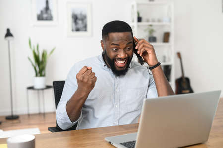 Successful Afro American freelancer talking on the phone, happy to achieve the goal. Young businessman looking at the screen of the laptop, winner, receiving good news in successful conversationの写真素材