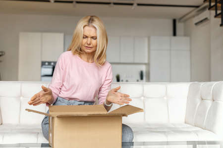 Disappointed middle-aged woman opens parcel carton box, unpacking it and feels sad about her wrong...