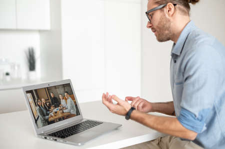 Intelligent young male entrepreneur in glasses sitting at the desk and presenting a project to a group of clients or colleagues via laptop, having a virtual online meetingの写真素材