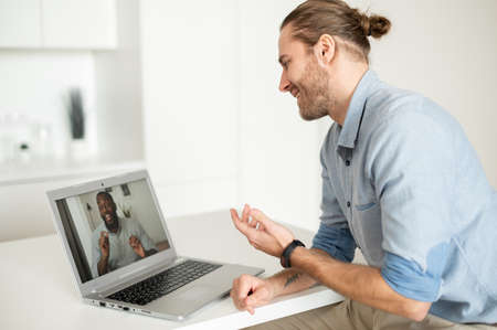Young handsome hipster guy sitting at the desk at the home office and talking to virtual African...
