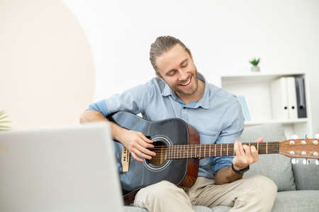 Attractive joyful bearded hipster man smiling and playing the guitar, sitting in the living room on...