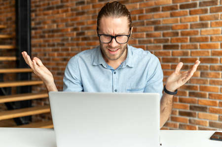 Young hipster guy in glasses sitting at the desk in front of a laptop and raising his hands up,...