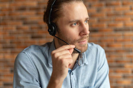 Close up of a millennial young businessman in a headset holding the microphone, attentive hipster customer service representative posing against the brick wall and responding to the customer inquiryの写真素材
