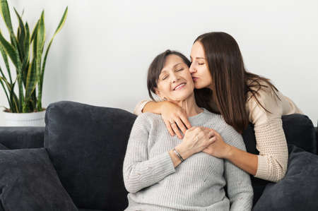 An adult daughter embracing and kissing an elder senior mother sitting on the sofa in cozy living...