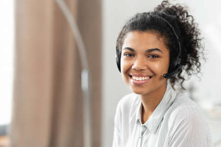 Headshot of friendly charming African American mixed-race woman in headset, smiling at camera working in the customer service department as call center operator, providing consultations on the phoneの写真素材