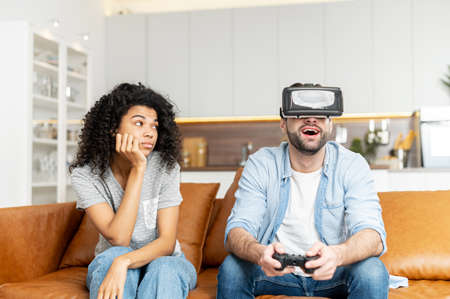 Amazed surprised guy wearing virtual reality glasses playing video game while her African girlfriend...