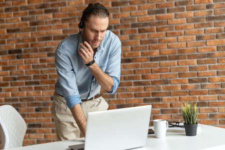 A guy wearing a headset deals with more than one task at the same time, male office employee stands...