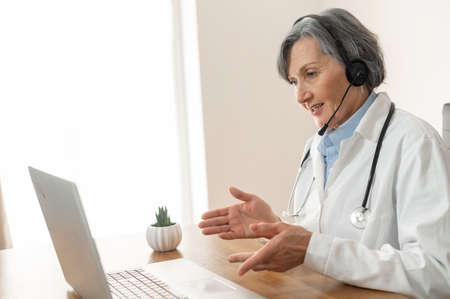 Senior old female doctor with stethoscope, wearing lab coat and headset, sitting in the office and...