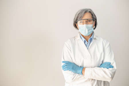 Senior female doctor medical robe, protective goggles, face mask, and latex gloves, crossing arms and posing isolated over gray background, fully equipped health professional working during pandemicの写真素材