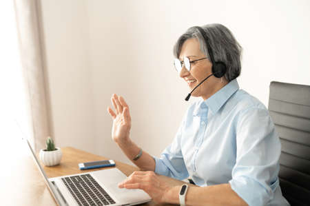 Mature female entrepreneur in glasses and a headset is sitting at the desk in home office and waving at the laptop screen, participating in an online virtual conference, chatting with teammatesの写真素材